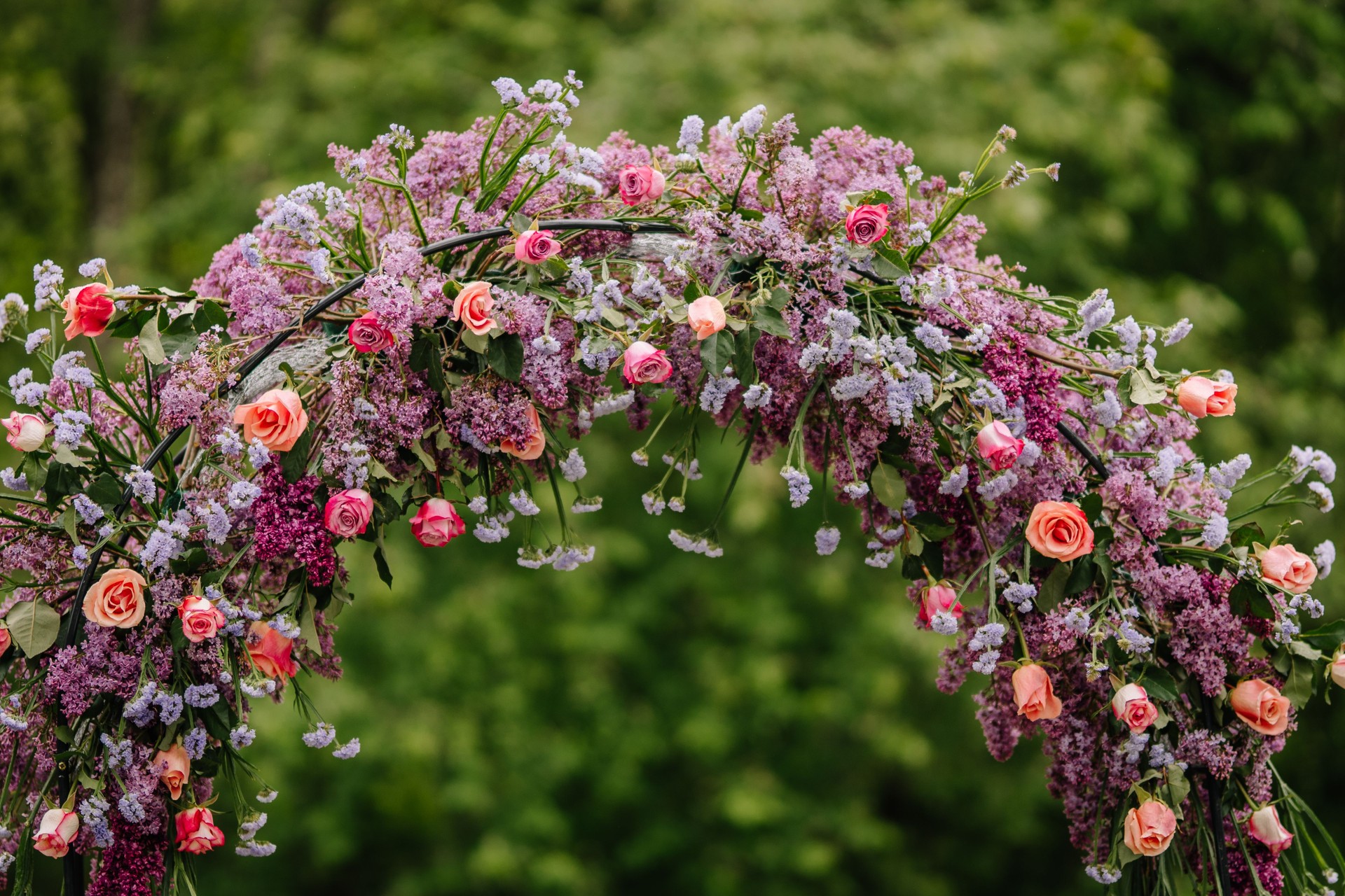 Close-up of a lush wedding floral arch made of lilacs, roses, and purple blossoms, with green foliage and soft natural background.